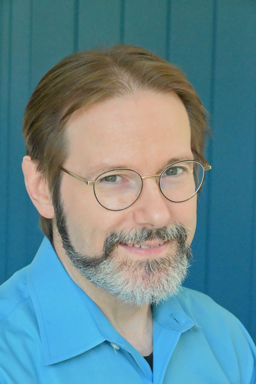 A headshot of a man wearing glasses and a blue shirt. He has brown hair and a darker mustache and beard which is greying.