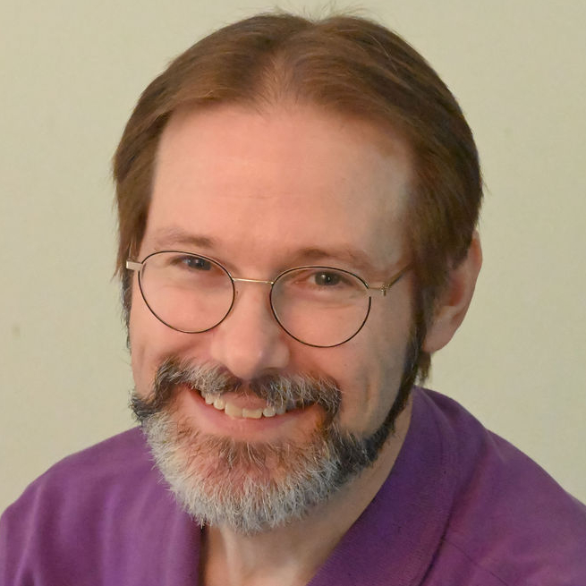 Headshot of a man wearing glasses and a purple shirt. His hair is brown and his beard and mustache are darker, but greying.